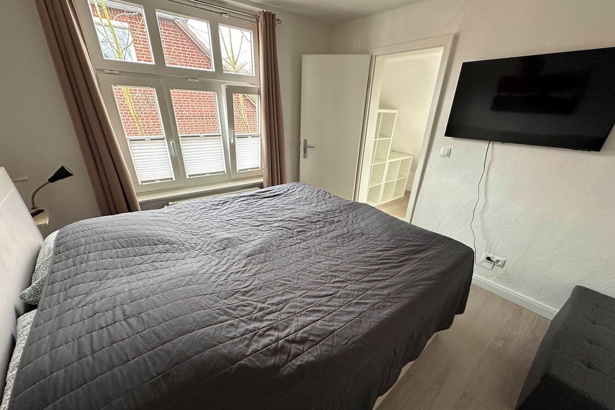 Ein Schlafzimmer mit einem großen Bett, einem an der Wand montierten Fernseher und einem Fenster mit Vorhängen.