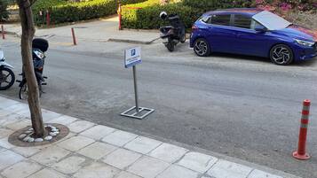 A parking area with a blue car, a scooter, and a motorcycle. There are potted plants and a tree on the sidewalk.