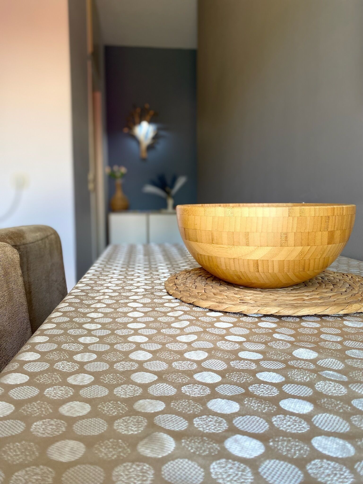 A wooden bowl on a woven placemat on a table with a patterned tablecloth.