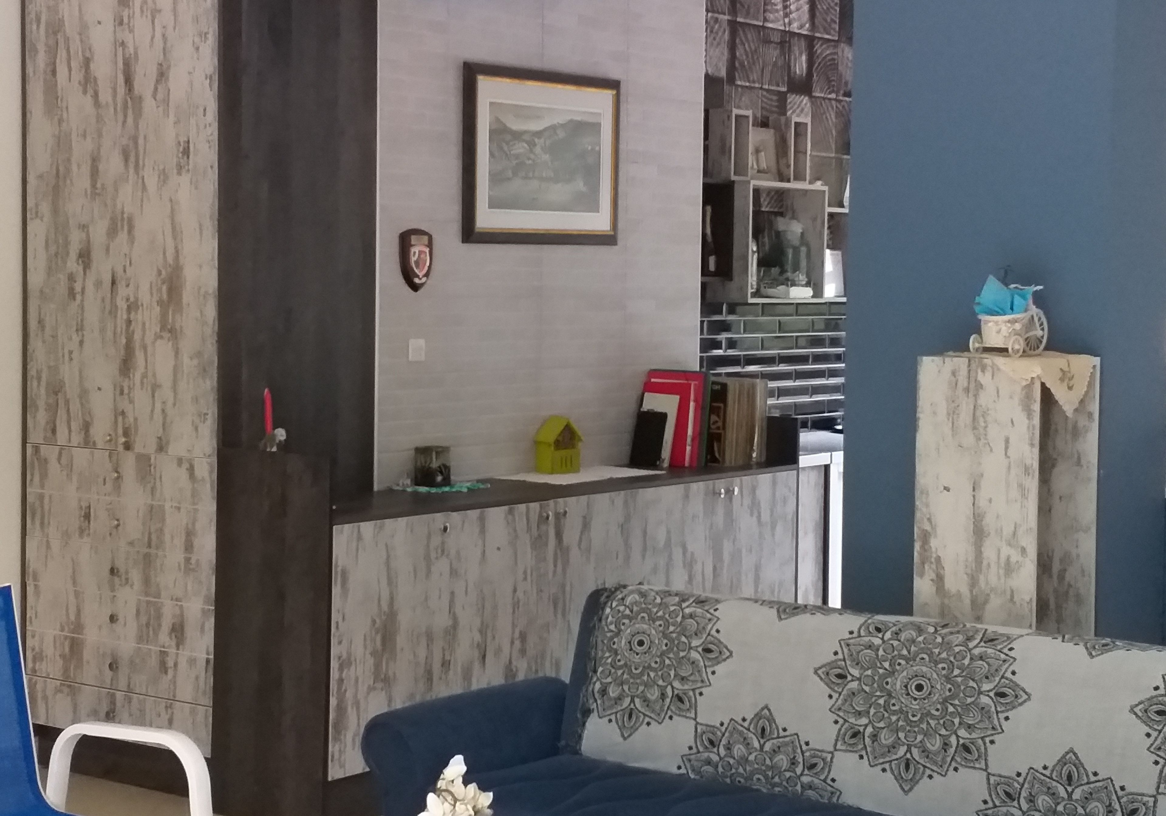 Uma sala de estar com um sofá azul, um armário de madeira, um quadro pendurado na parede e uma pequena estante com objetos decorativos.