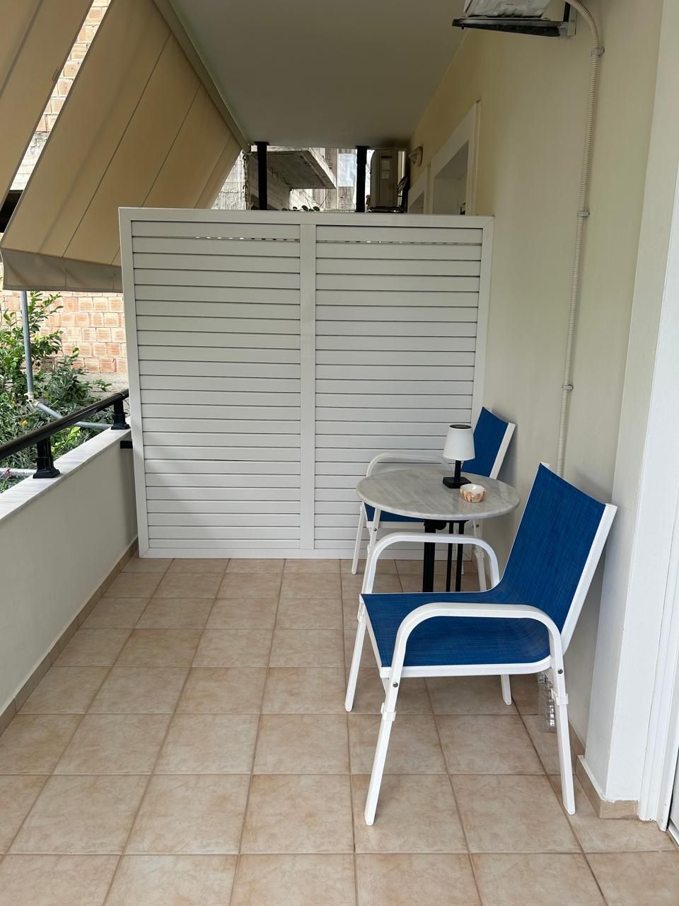 A balcony with a table and two chairs, a tiled floor, and a white wall with a window.