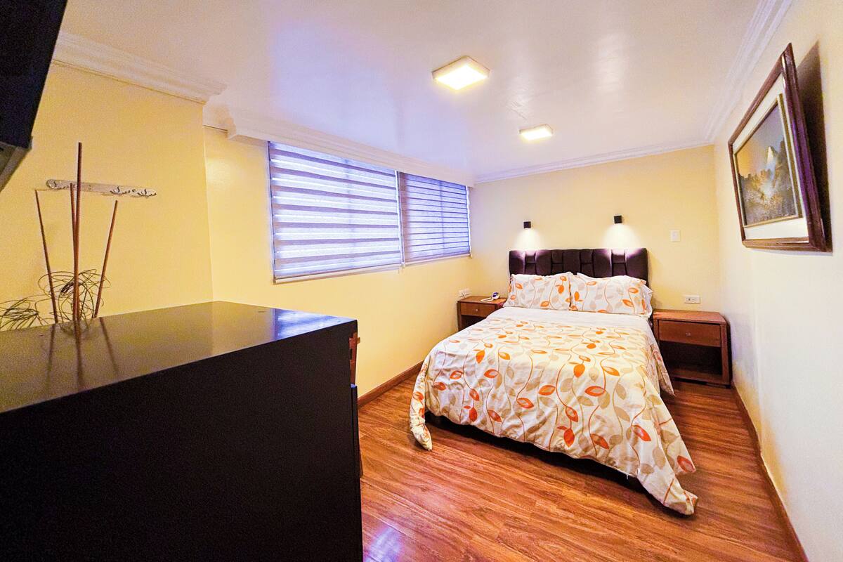 Quarto com cama, mesas de cabeceira, cômoda e janela com persianas.