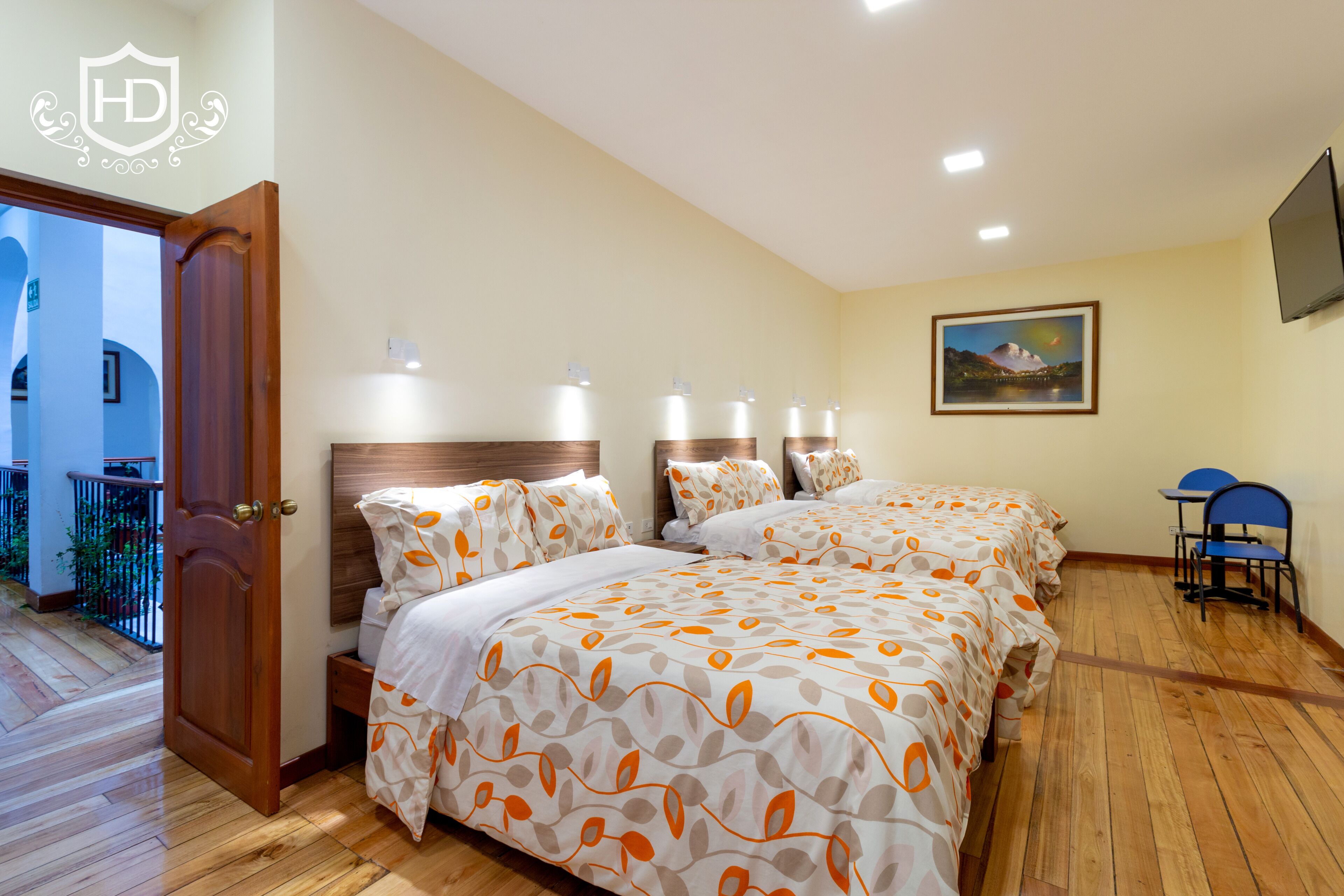 A hotel room with a wooden floor, two beds with matching headboards, a painting on the wall, and a TV mounted above a desk.