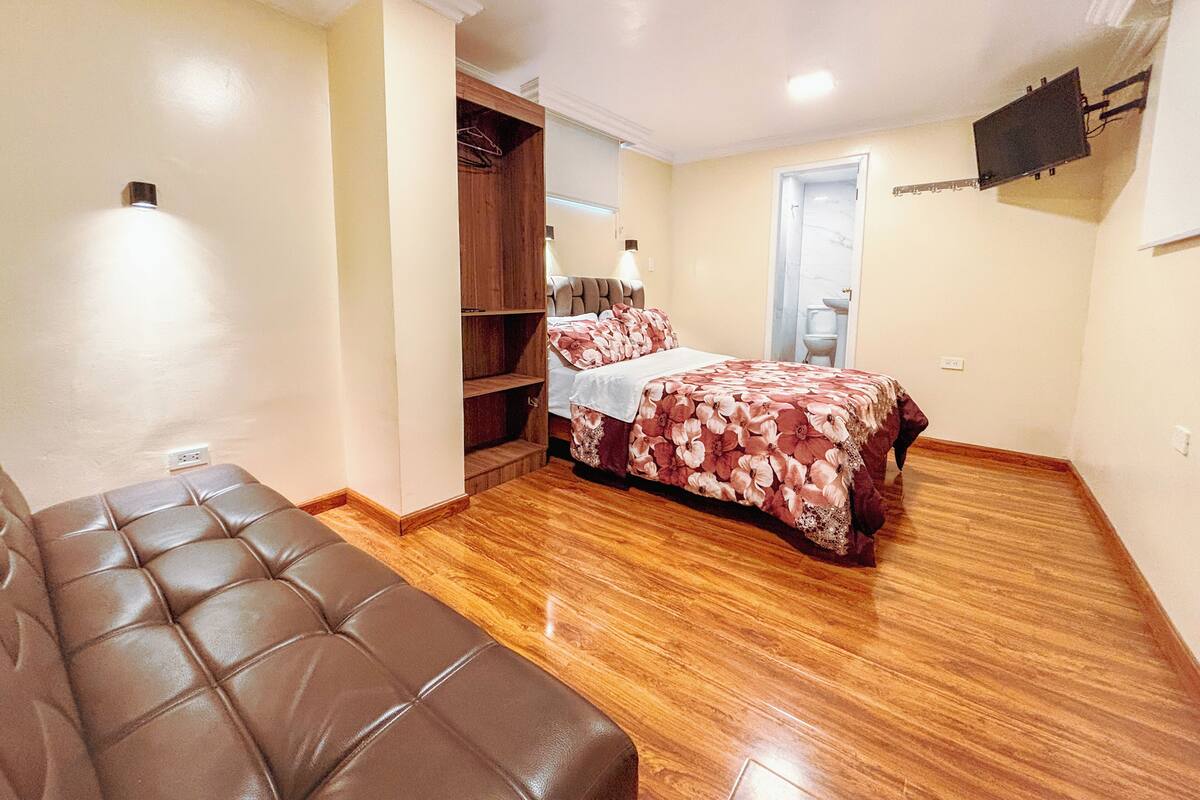 Quarto com cama, guarda-roupa de madeira, sofá de couro marrom e televisão fixada na parede.