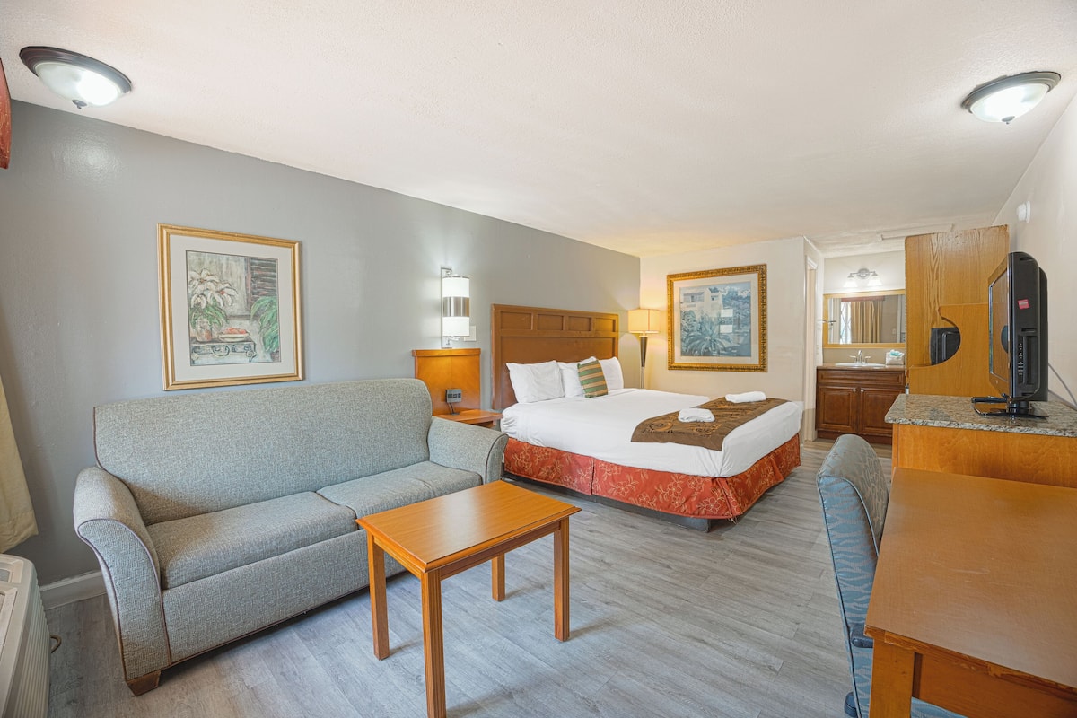 Ein Hotelzimmer mit Bett, einer Couch, einem kleinen Tisch, einem Fernseher und einem Badezimmer mit Spiegel und Waschbecken.