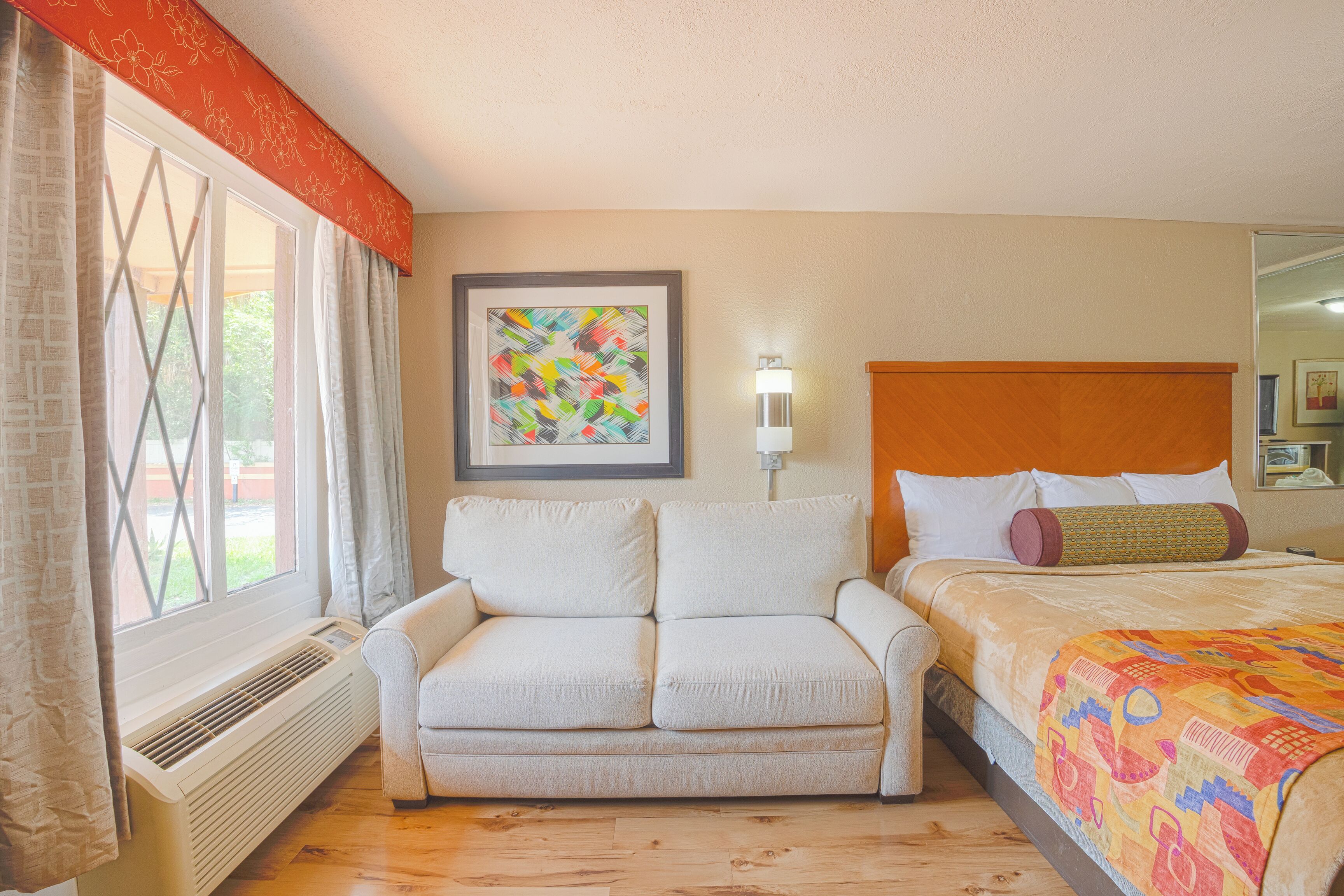 A hotel room with a white sofa, a bed with a colorful quilt, a framed abstract painting, a wall-mounted lamp, and a window with curtains.
