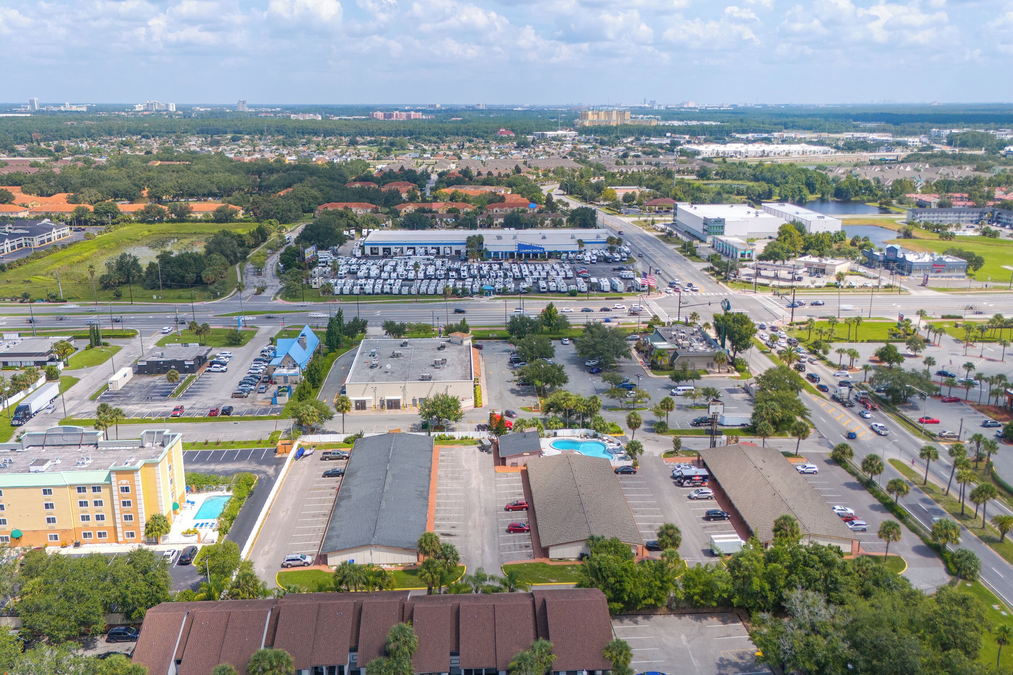 An aerial view of a commercial area with a large parking lot, a swimming pool, and a hotel with multiple floors and a flat roof.