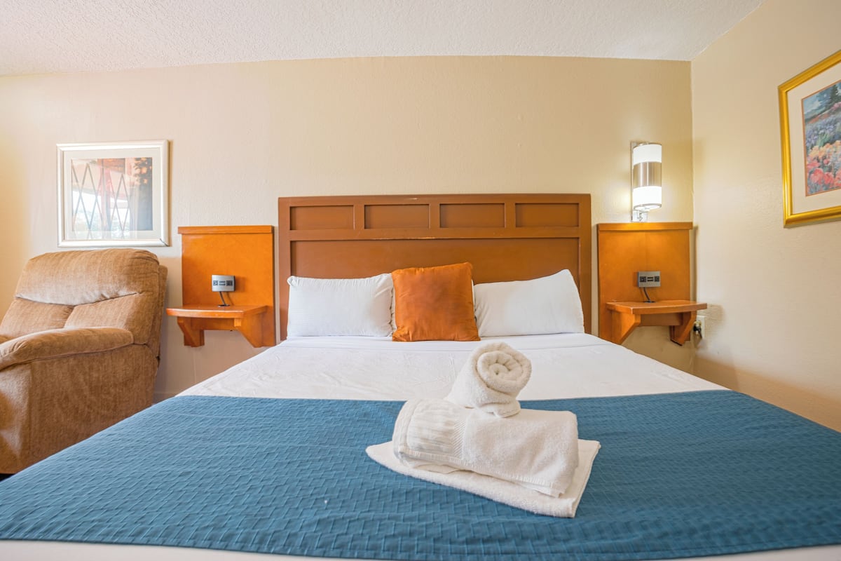 Ein Hotelzimmer mit einem großen Bett, einem Sessel, zwei Nachttischen mit Lampen und einem gerahmten Bild an der Wand.