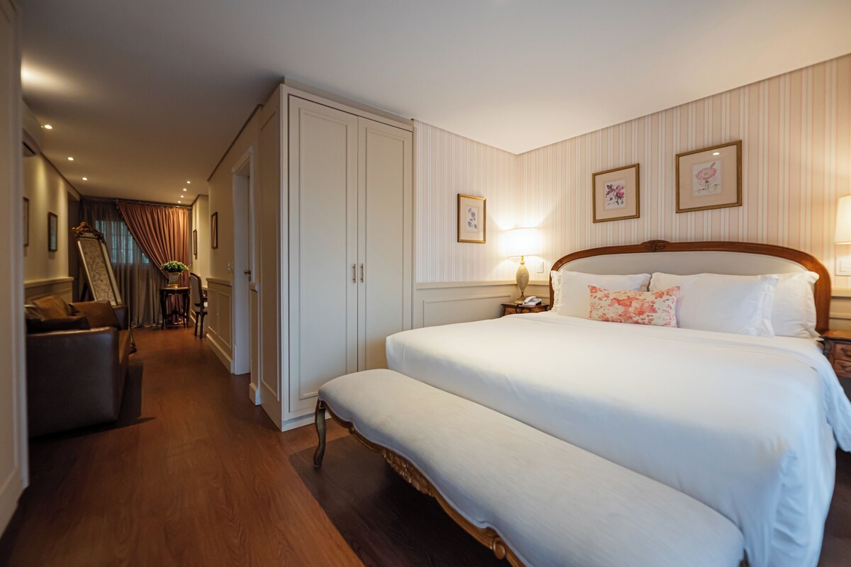 A hotel room with a large bed, a bench, a desk, a chair, a wardrobe, and a mirror.