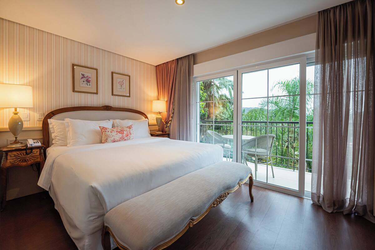 Habitación de hotel con una cama grande, dos lámparas de noche, una mesita de noche y un balcón con vistas a zonas verdes.