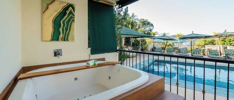 Un área de spa con una bañera grande, un banco de madera y vistas a una piscina con sillones y sombrillas.