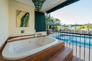 A spa area with a large bathtub, a wooden bench, and a view of a pool with lounge chairs and umbrellas.