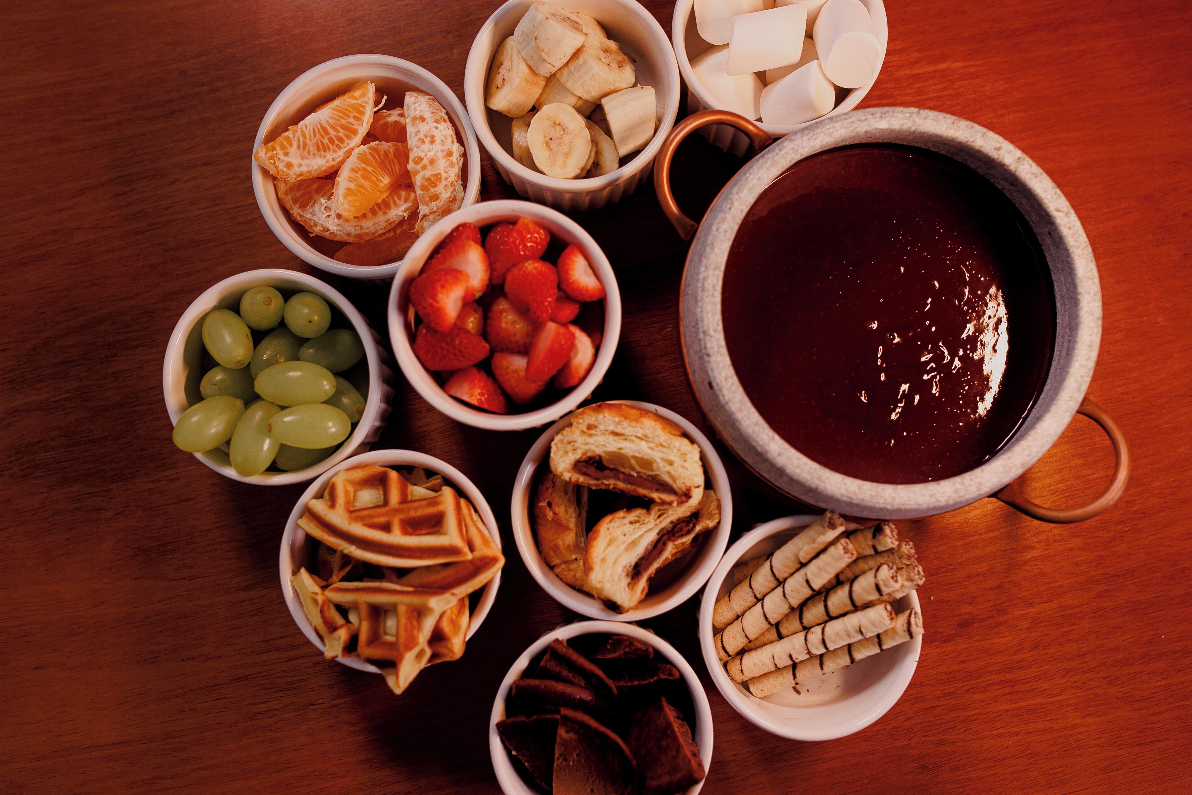 Diversos alimentos, entre ellos frutas, pan y un tazón grande con un líquido oscuro, probablemente una bebida, dispuestos sobre una mesa de madera.