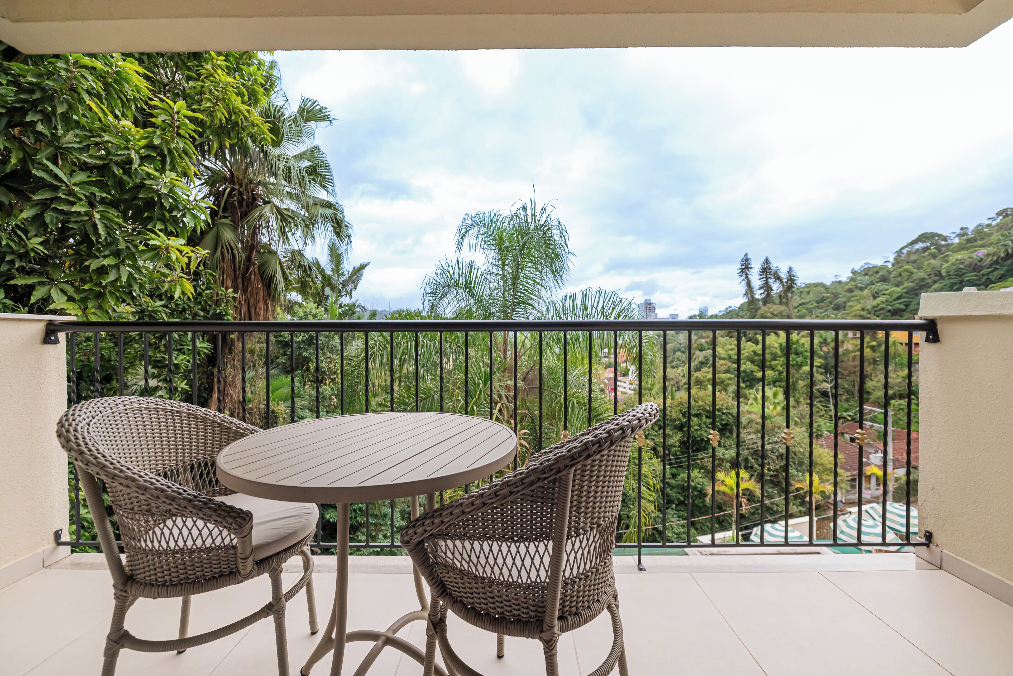 Un balcón con sillas de mimbre y una mesa, con vista a un paisaje verde y exuberante con árboles y edificios a lo lejos.