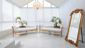 Una habitación con techo blanco, una araña, dos bancos blancos acolchados, plantas en macetas y un espejo grande y ornamentado.