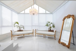 A room with a white ceiling, a chandelier, two white cushioned benches, potted plants, and a large ornate mirror.