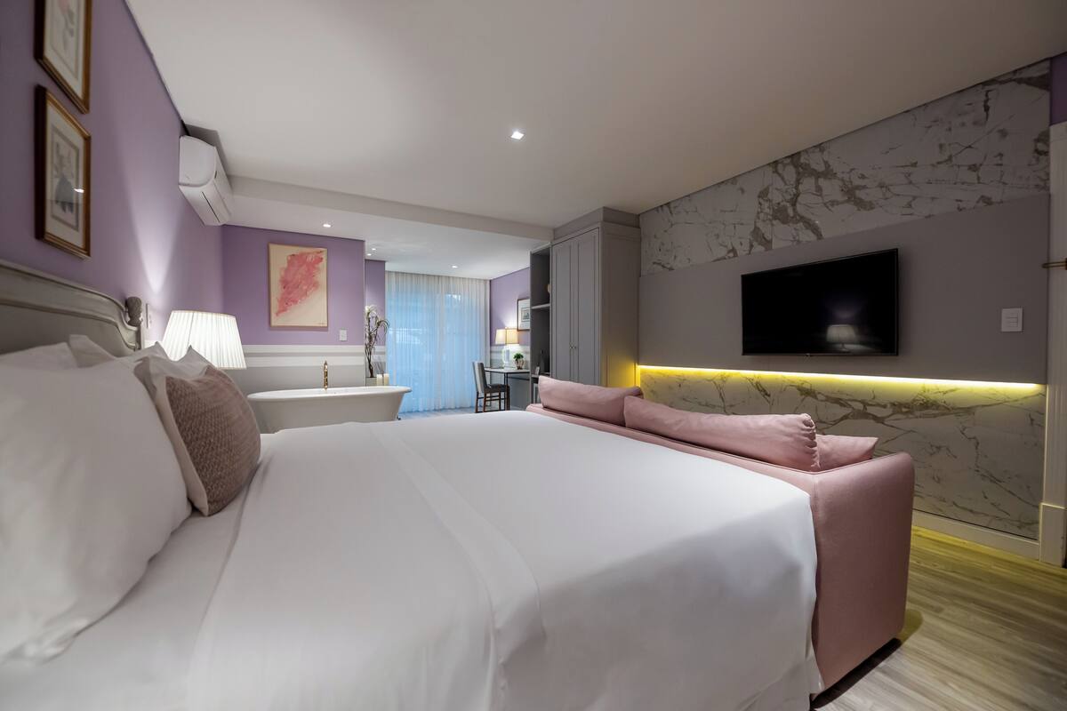 Una habitación de hotel moderna con una cama grande, un televisor montado en la pared y un baño visible al fondo.