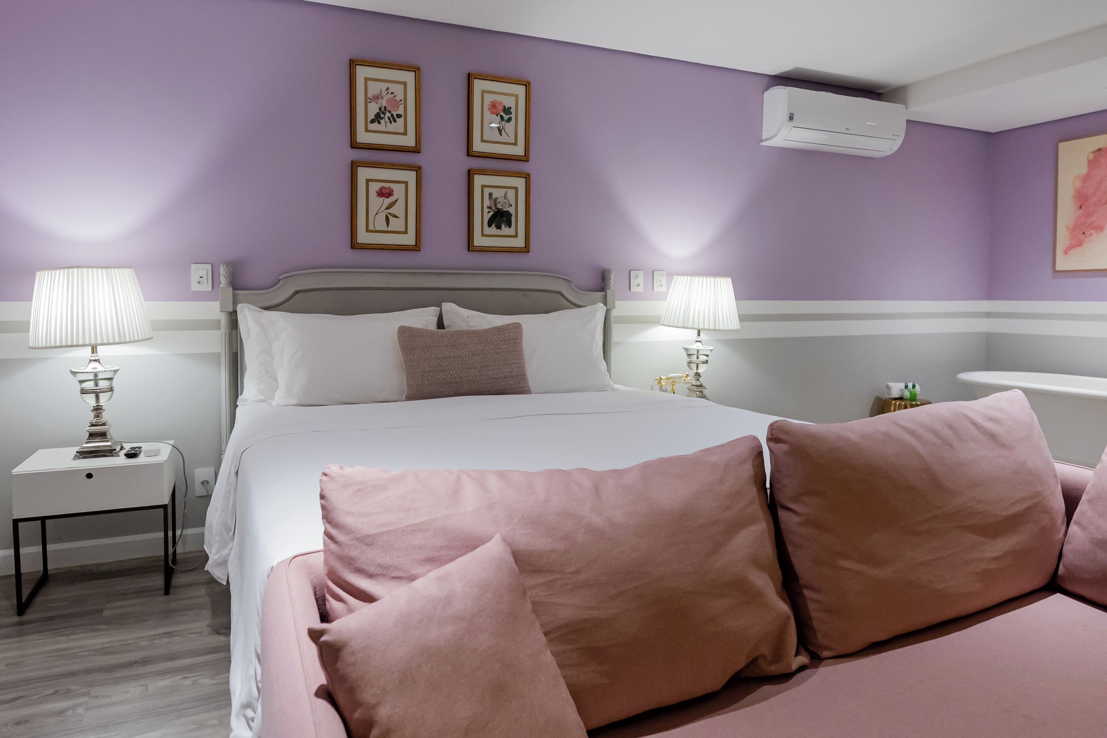 Un dormitorio con cama, dos mesitas de noche con lámparas, una mesita auxiliar, un aire acondicionado de pared y cuadros con motivos florales.