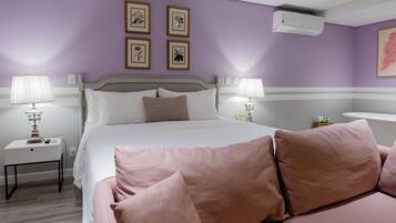 Un dormitorio con cama, dos mesitas de noche con lámparas, una mesita auxiliar, un aire acondicionado de pared y cuadros con motivos florales.