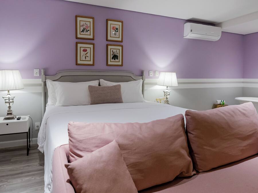 Un dormitorio con cama, dos mesitas de noche con lámparas, una mesita auxiliar, un aire acondicionado de pared y cuadros con motivos florales.