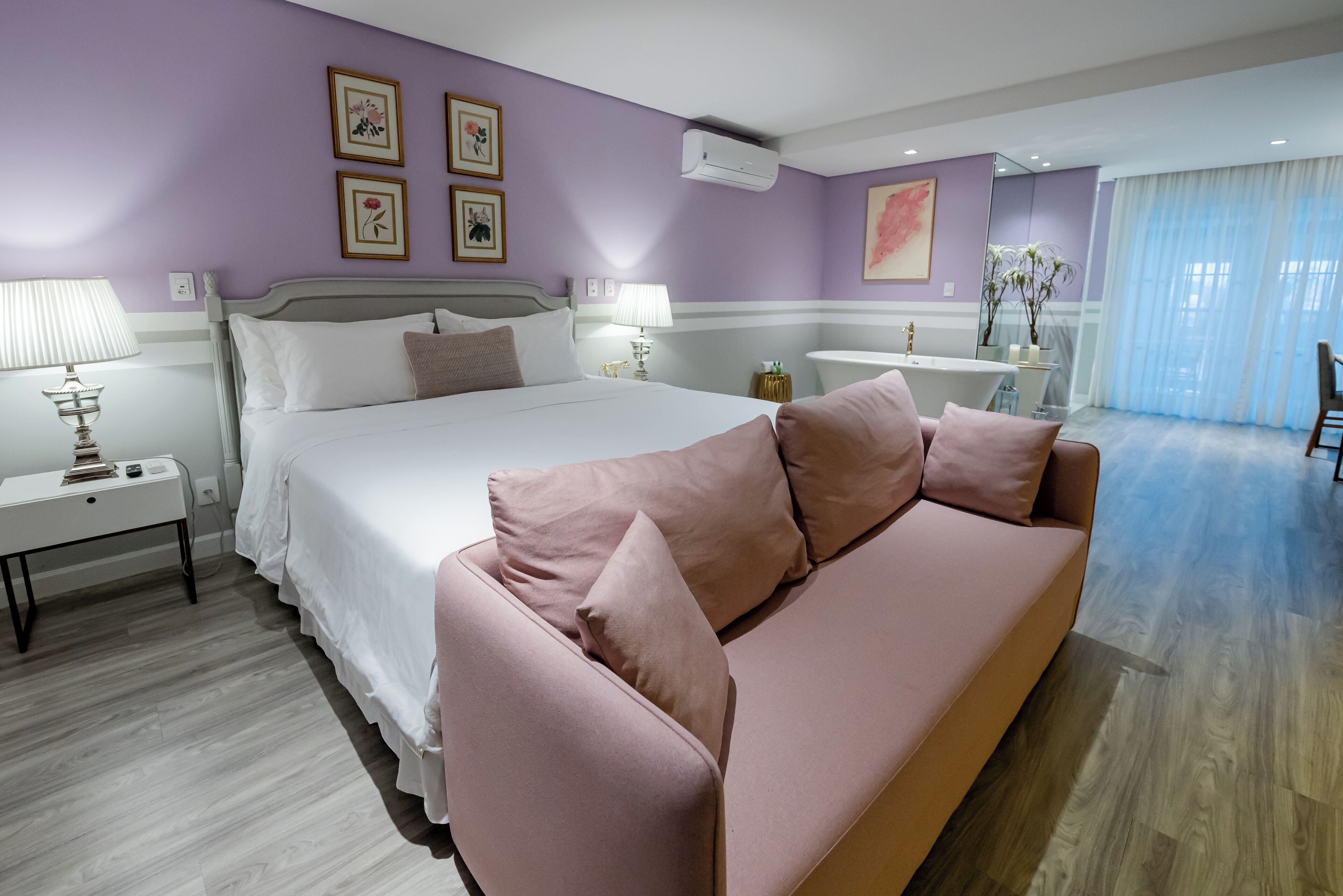 Un dormitorio con una cama, un sofá rosa, una mesita de noche con lámpara y un baño al fondo.