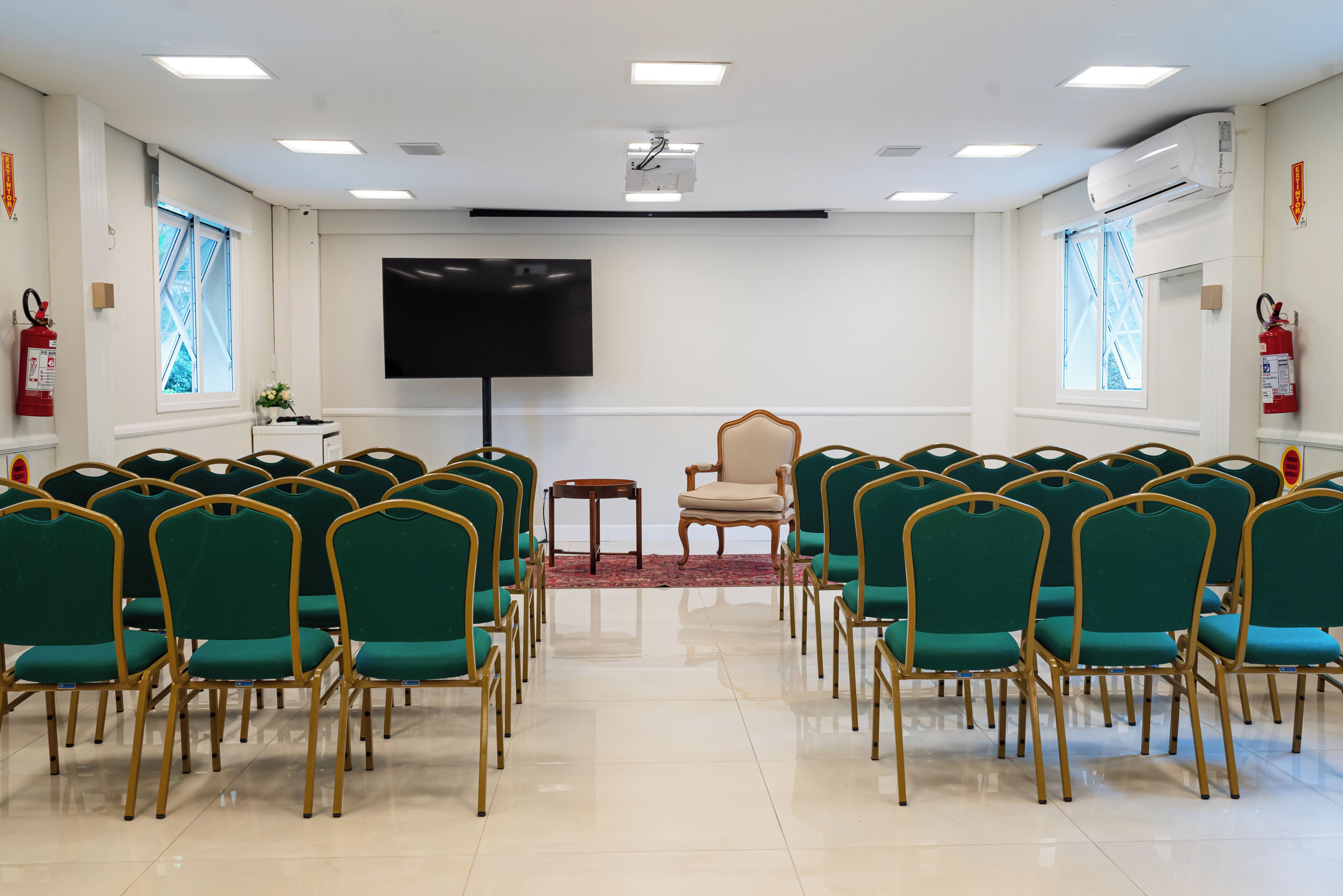 Un salón de conferencias con filas de sillas verdes, una pantalla de proyector y un extintor.