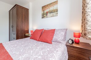 Un dormitori amb un llit, almoinos vermells, una nitòria amb una làmpada, un rellotge i un quadre a la paret.