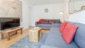 Un salon avec un téléviseur posé sur un meuble en bois, un canapé gris agrémenté de coussins rouges, une petite table en bois et un grand tableau abstrait accroché au mur.