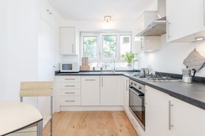 A modern kitchen with white cabinets, a black countertop, a microwave, a stove, and a window with a view of trees. - New Apartment Liverpool Street Brick Lane (London)