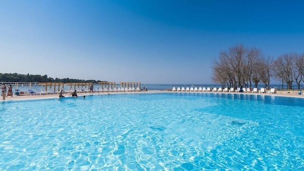 A large outdoor swimming pool with clear blue water, surrounded by white lounge chairs and a wooden pergola.