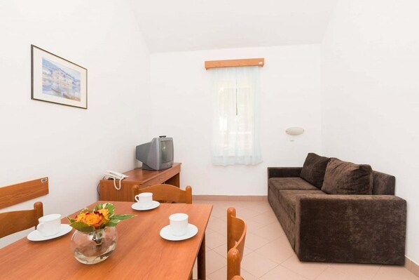 A small, clean room with a wooden dining table set for two, a brown sofa, a television, and a framed picture on the wall. - Polynesia Apartments Plava Laguna (Umag)