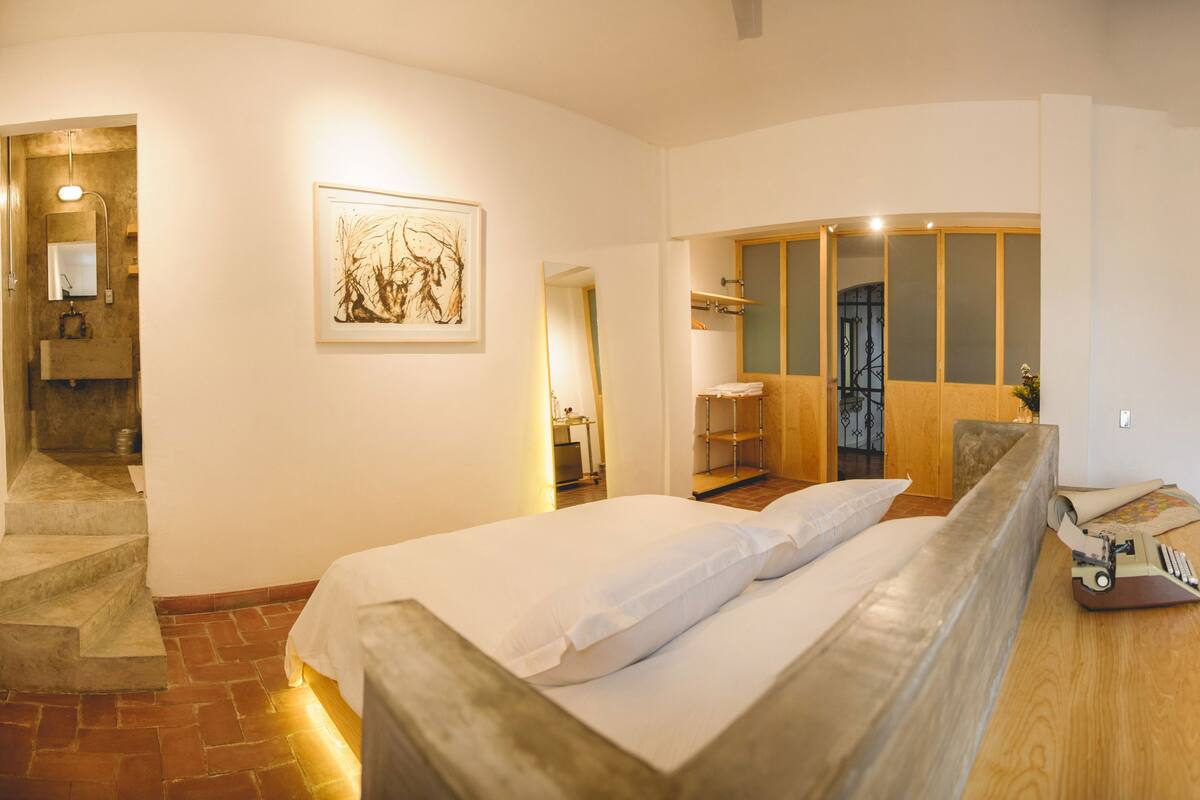 A bedroom with a bed, a wooden bench, a painting, a bathroom with a shower, and a staircase.