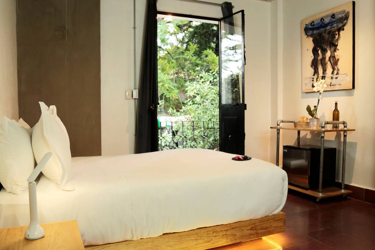A modern bedroom with a large bed, a bedside table, a nightstand, and a view of greenery through an open door.