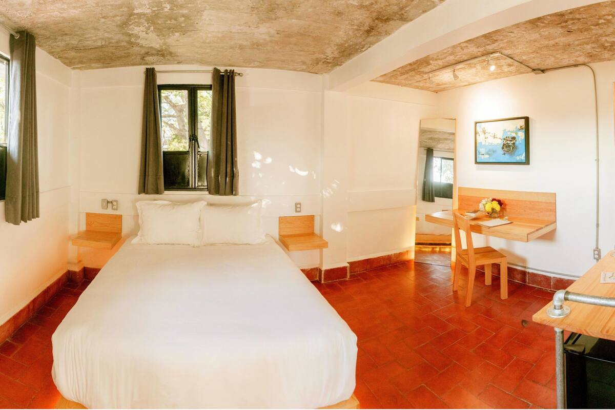A bedroom with a large bed, a desk, and a chair. There are curtains on the windows, and a painting on the wall.