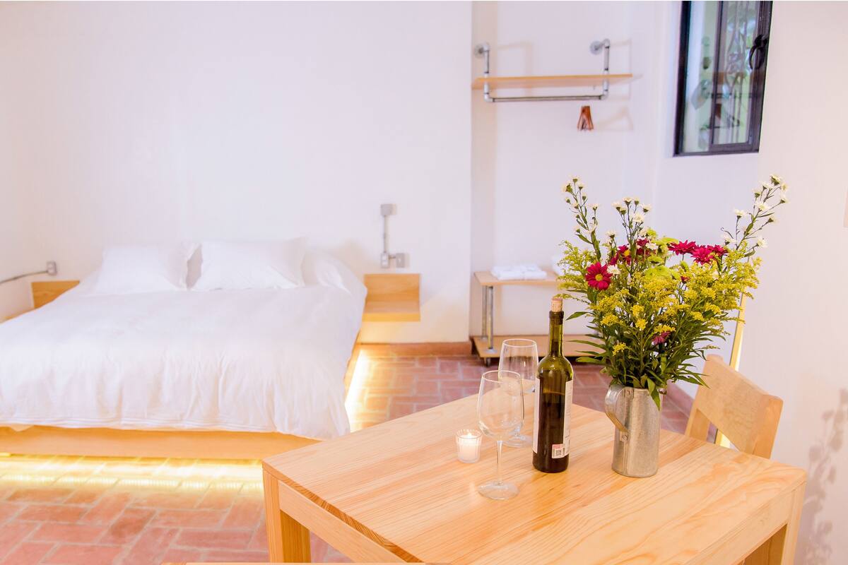 A hotel room with a bed, a wooden table with a vase of flowers, two wine glasses, and a bottle of wine.