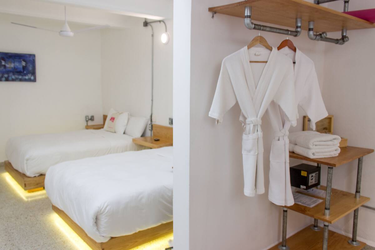 A hotel room with two beds, a wooden nightstand, a robe hanging on a rack, and a shelf with towels.