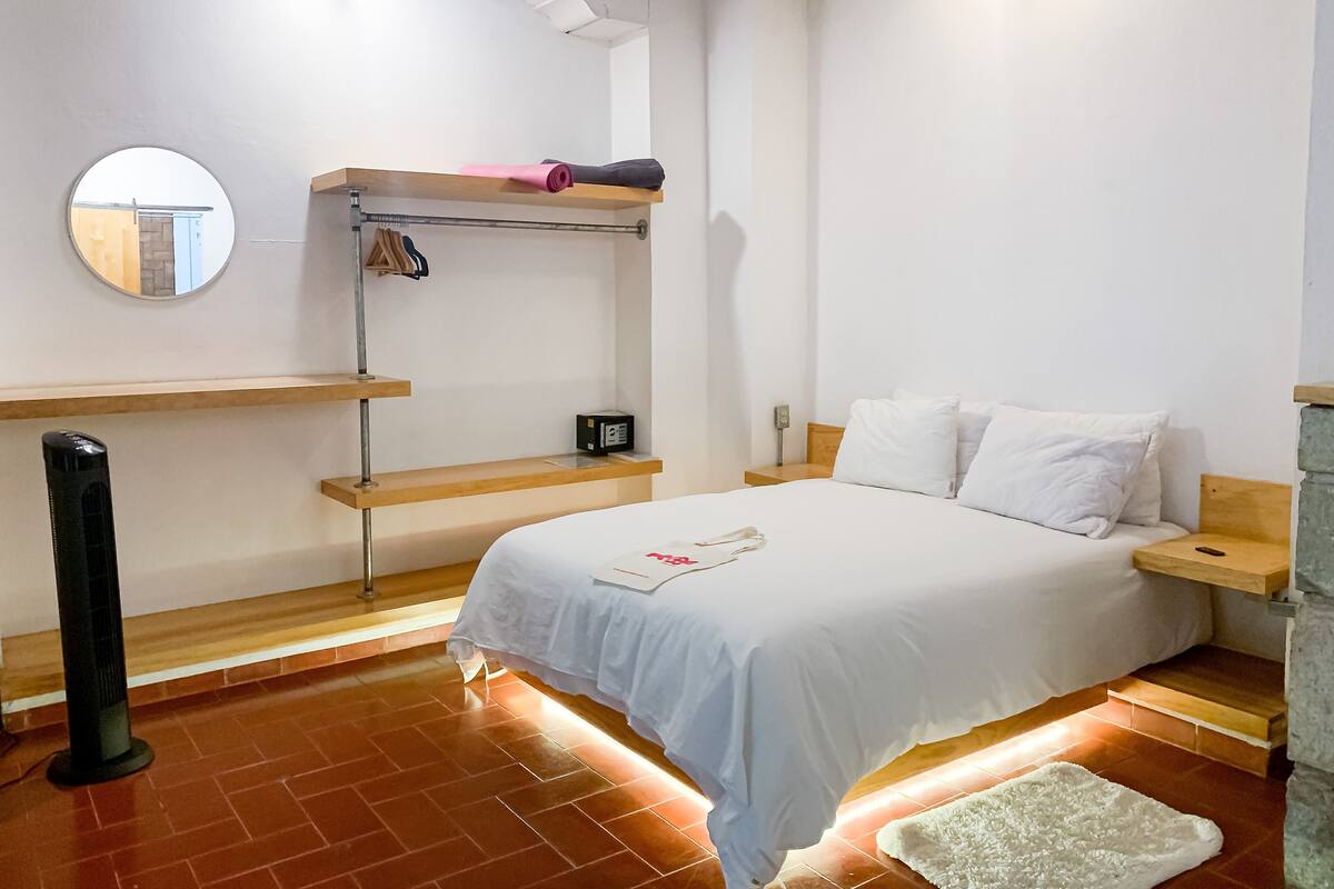 A bedroom with a bed, a bedside table, a mirror, a shelf with clothes, and a heater.