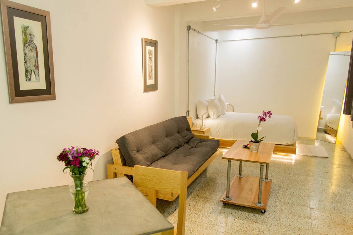 A room with a sofa, a small table with a vase, and a bed with a nightstand.