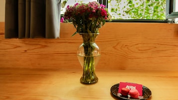 Una mesa de madera con un florero de vidrio transparente que contiene flores rosas, un plato negro con dos macarons rojos y una tarjeta roja con la palabra "AGRE" parcialmente visible.