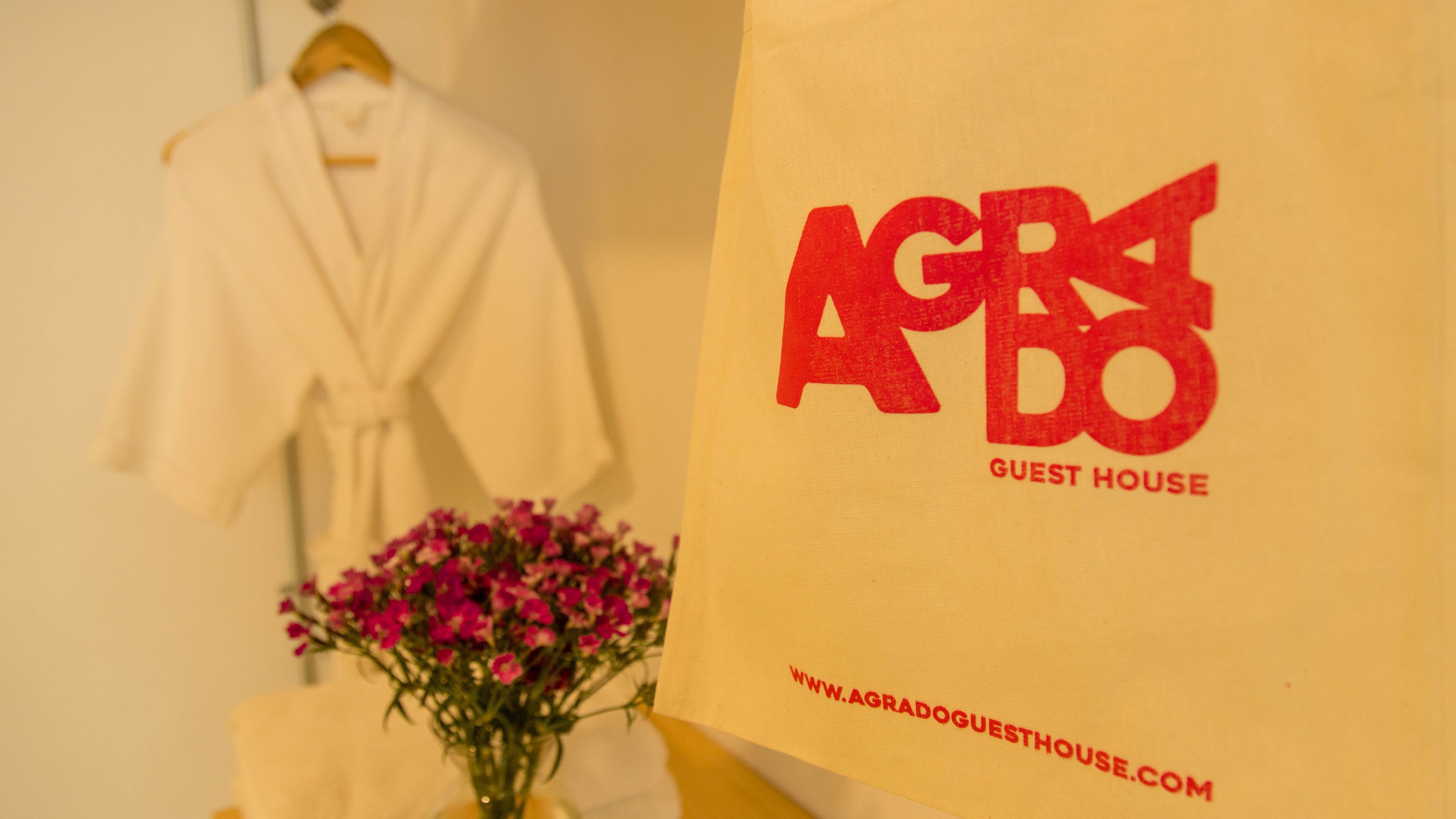 A white robe hanging on a wooden hanger, a vase with pink flowers, and a yellow bag with red text reading "AGBO GUEST HOUSE" and a website address.