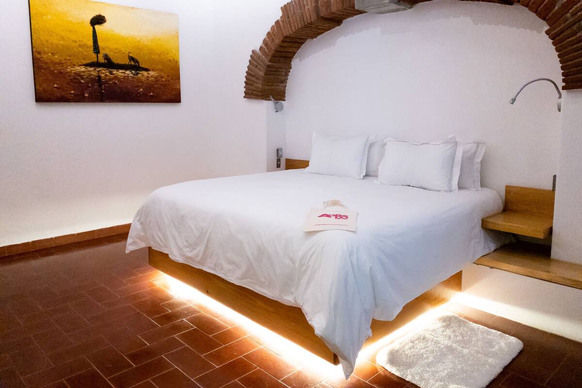 A bedroom with a bed, a white pillow, a wooden bench, a white rug, a painting, and a brick archway.
