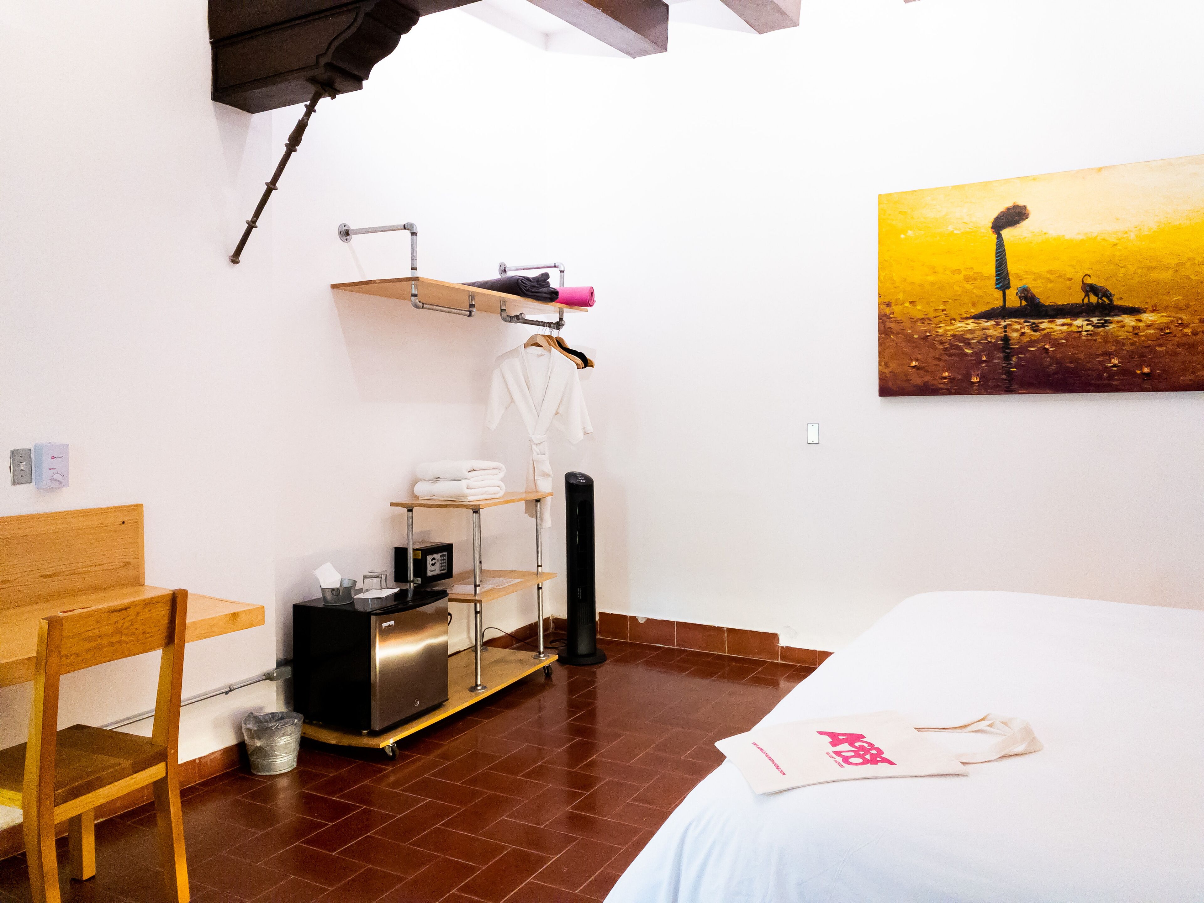 Una habitación de hotel con una cama, una silla de madera, una mesita con un radio, un estante con toallas y una bata, y un cuadro en la pared.