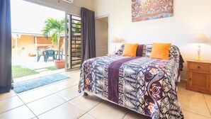 A bedroom with a bed, a nightstand, a lamp, and a view of the outside. - Au Regina (Arue)