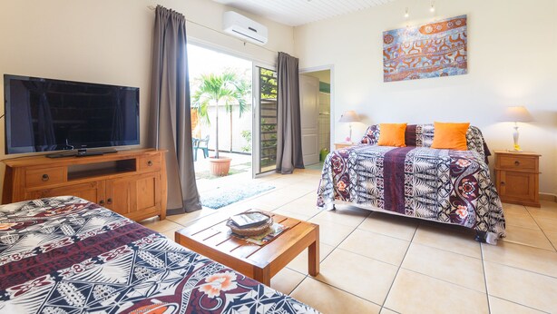 A bedroom with a bed, a television, a wooden cabinet, and a window with curtains. - Au Regina (Arue)