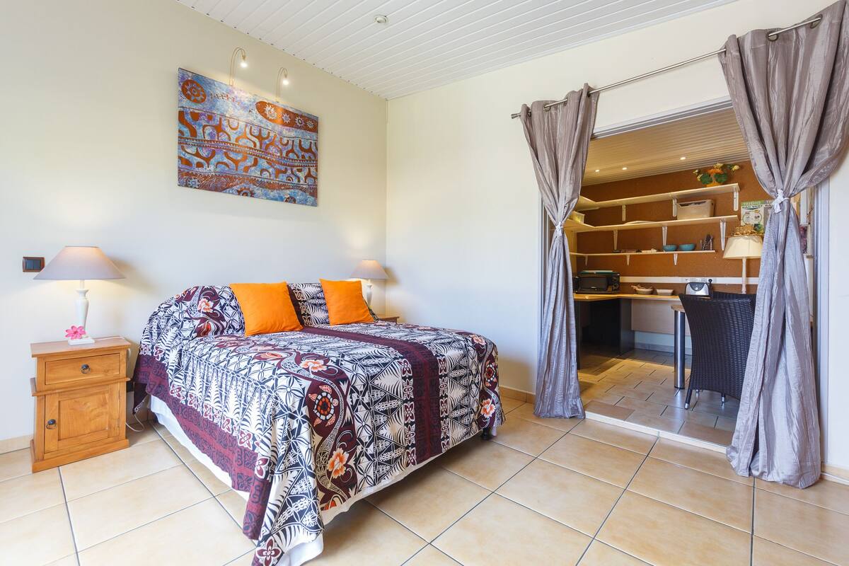 A bedroom with a bed, nightstands, a painting, and a kitchenette in the background.