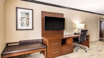 A hotel room with a wooden desk, a flat-screen TV, a bench, a desk chair, and a lamp.