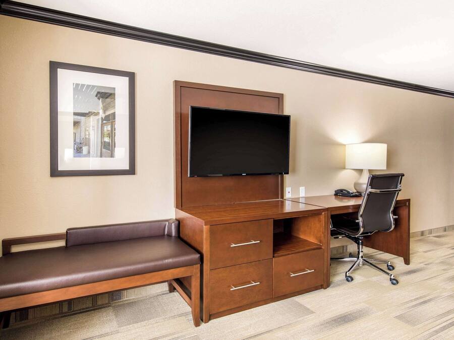 A hotel room with a wooden desk, a flat-screen TV, a bench, a desk chair, and a lamp.