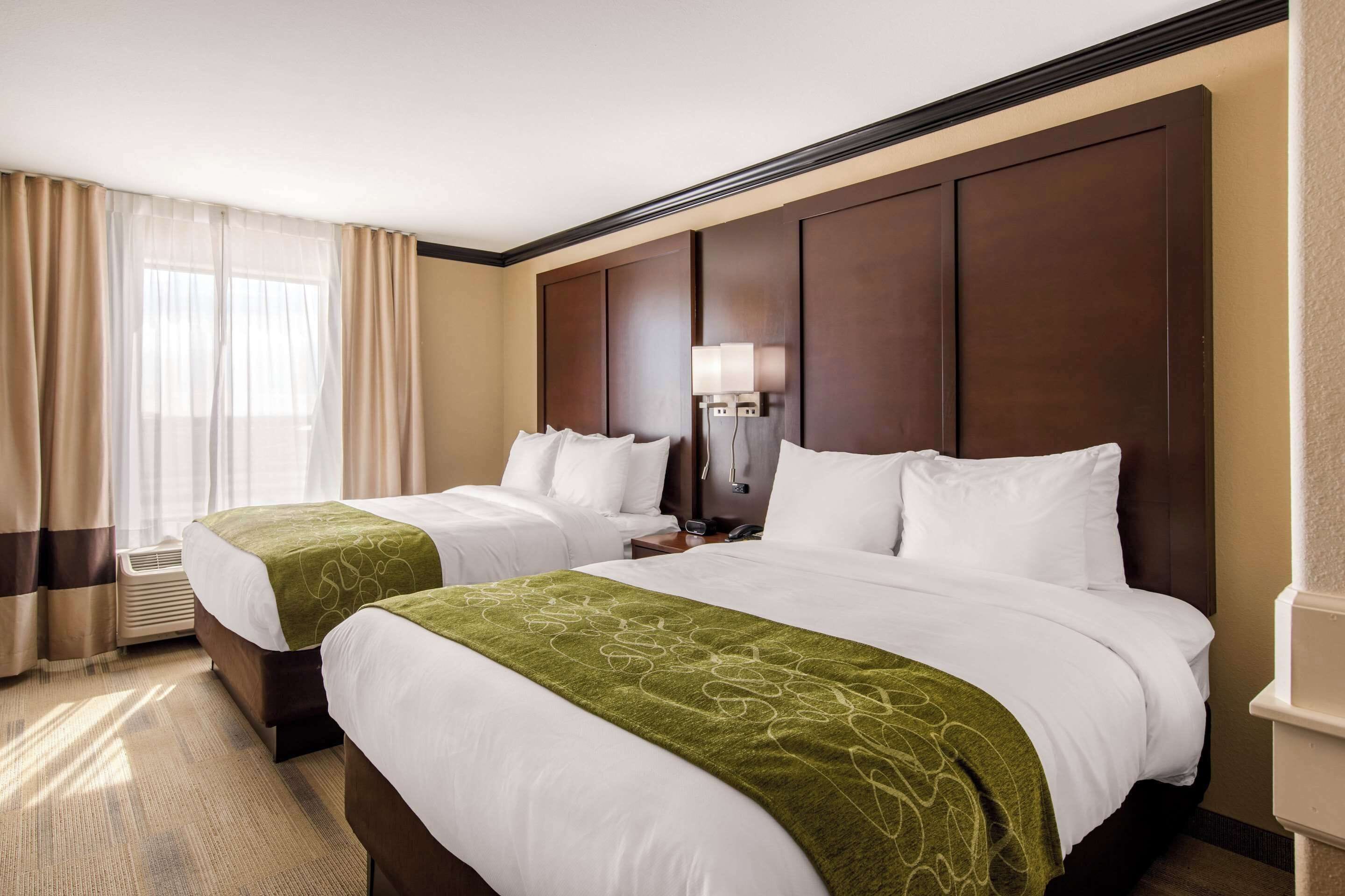 A hotel room with two beds, a large window with curtains, a wall-mounted lamp, and a nightstand.