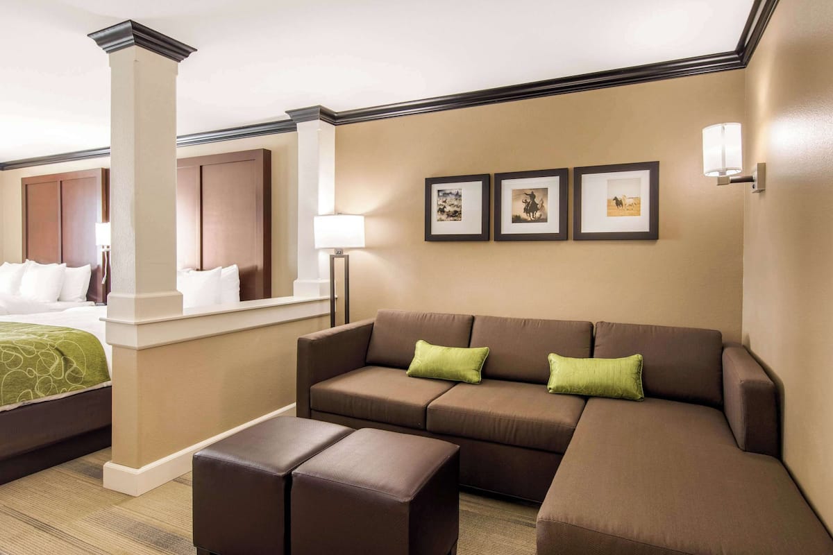 A hotel room with a large sectional sofa, a small table, and a bed with a green bedspread. There are framed pictures on the wall and a lamp on a side table.