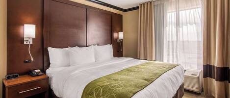 Une chambre d’hôtel avec un grand lit, deux tables de chevet, une petite table, une lampe fixée au mur et une fenêtre avec des rideaux.